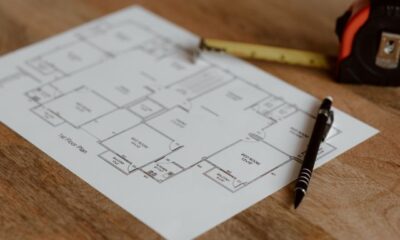 A residential floor plan printed on white paper, resting on a wooden table, with a yellow tape measure and a black pen placed on top of it.