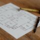 A residential floor plan printed on white paper, resting on a wooden table, with a yellow tape measure and a black pen placed on top of it.