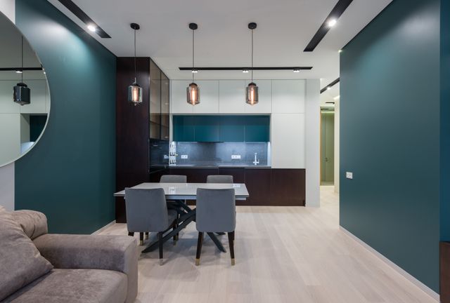 A modern small living room dining room combo featuring teal accent walls, a grey dining set under three glass pendant lights, and a sleek kitchen in the background.