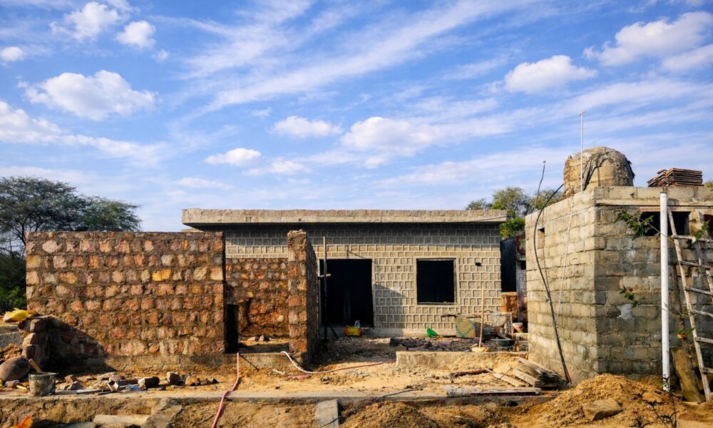 A partially built rural house with stone and concrete block walls, an unfinished roof, and scattered construction materials under a bright blue sky with scattered clouds.