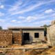 A partially built rural house with stone and concrete block walls, an unfinished roof, and scattered construction materials under a bright blue sky with scattered clouds.