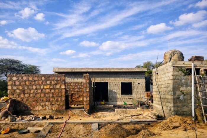 A partially built rural house with stone and concrete block walls, an unfinished roof, and scattered construction materials under a bright blue sky with scattered clouds.