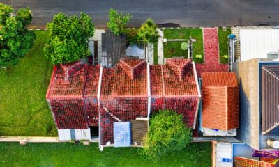 Aerial drone view of a red clay tile roof house surrounded by green trees, lawn, and a paved street