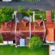 Aerial drone view of a red clay tile roof house surrounded by green trees, lawn, and a paved street