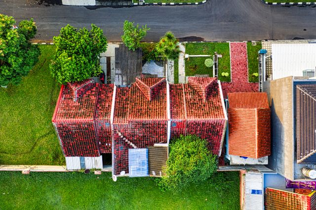Aerial drone view of a red clay tile roof house surrounded by green trees, lawn, and a paved street